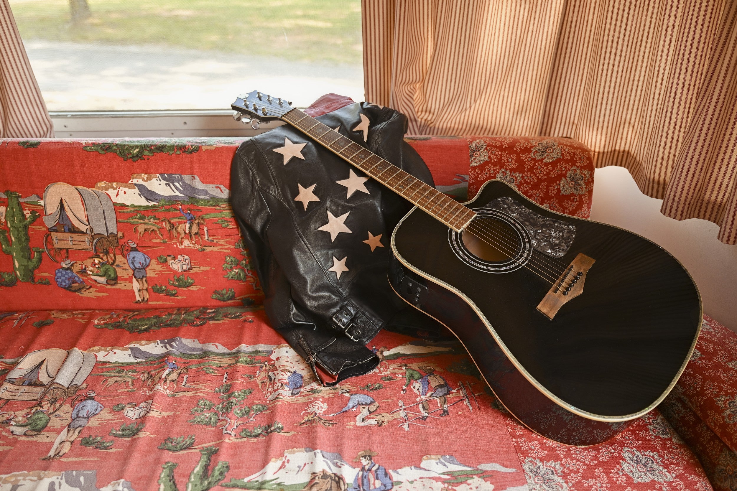 A black acoustic guitar leaning against a black leather jacket adorned with star patches, resting on a red vintage western-print couch with striped curtains in the background.