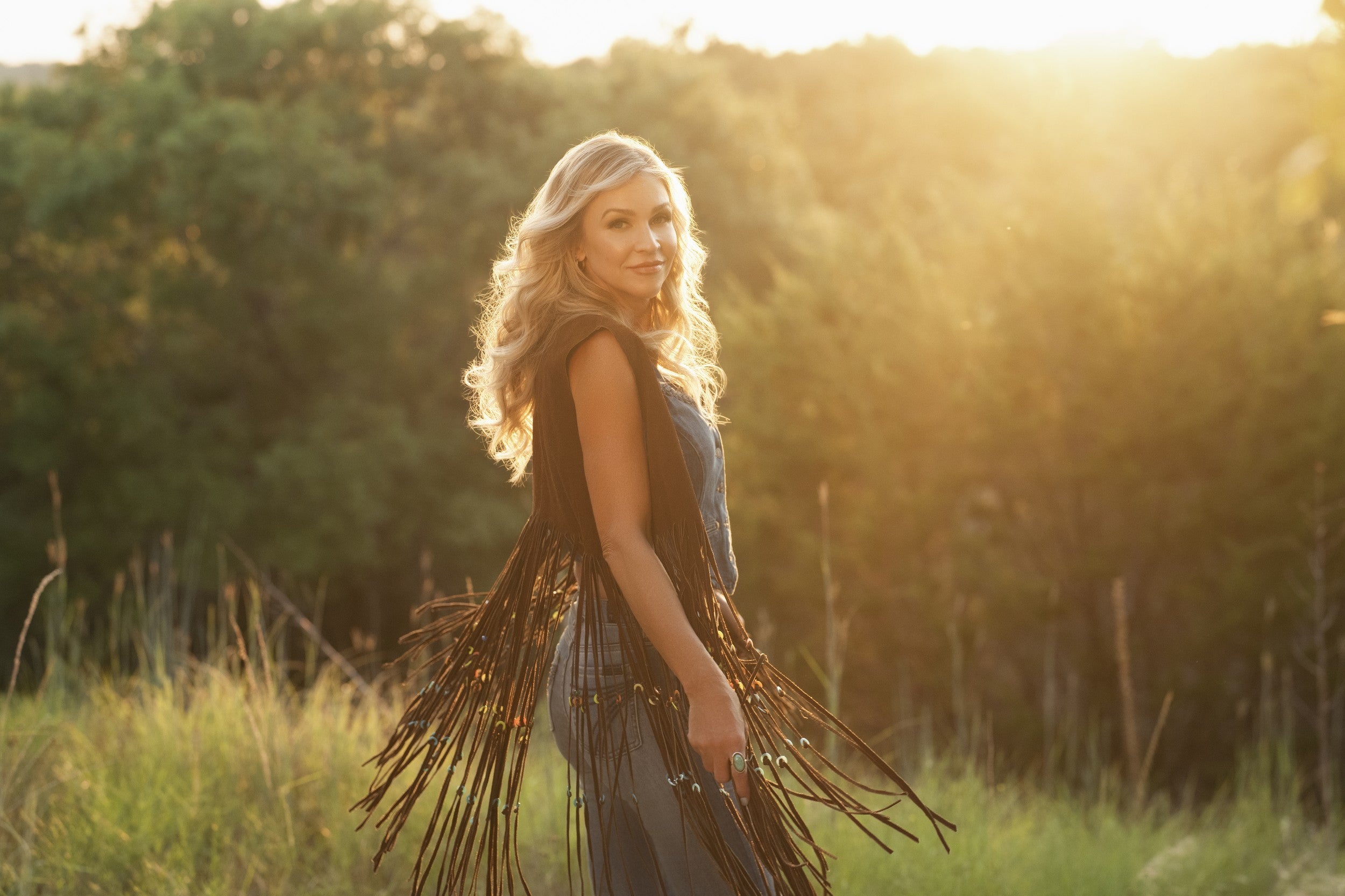 Tara Jennings standing in a golden field at sunset, looking back over her shoulder with a soft smile, wearing a long dark fringe vest over a sleeveless outfit, with tall grass and sunlit trees in the background.