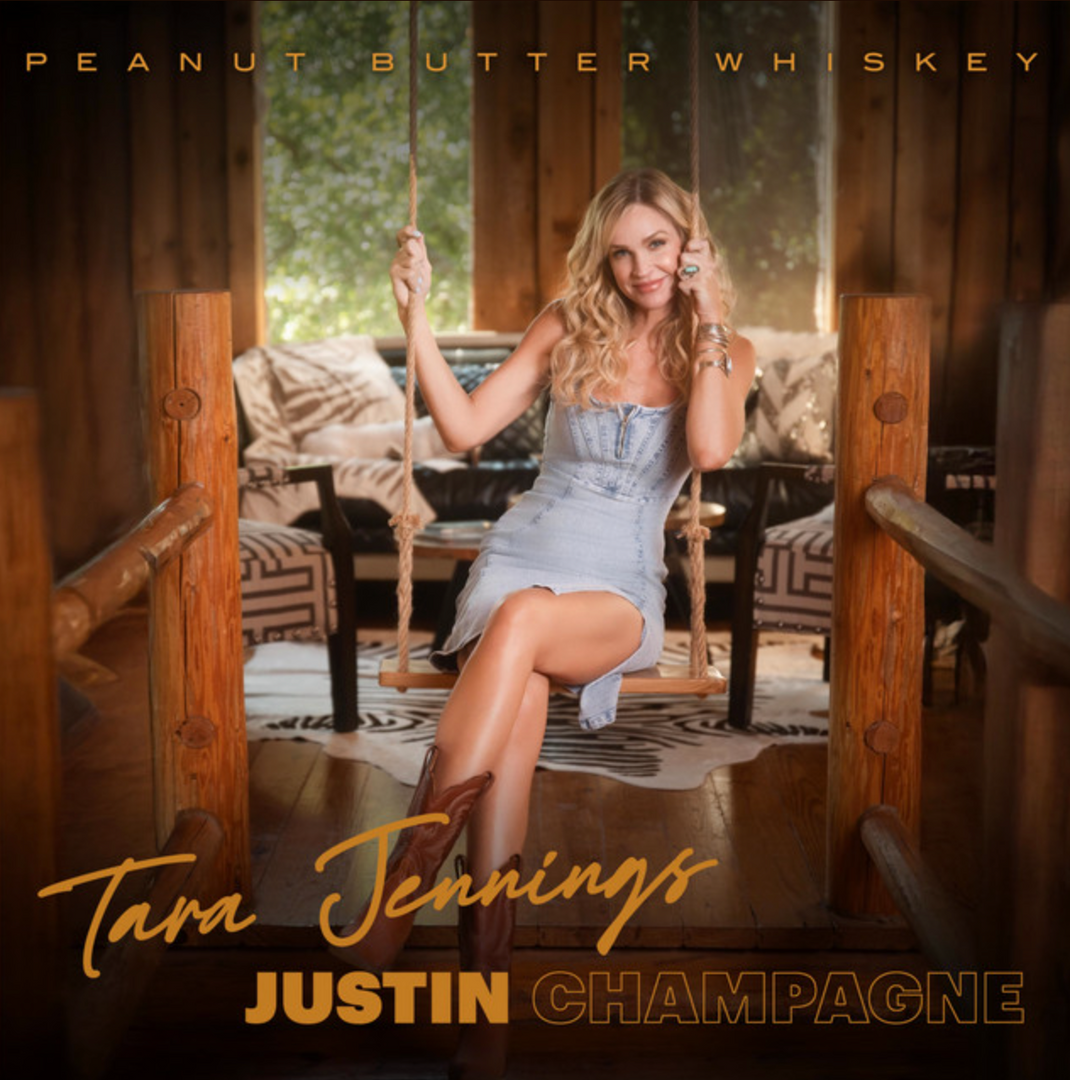 Tara Jennings sitting on a wooden swing, smiling, wearing a light blue denim strapless dress and brown cowboy boots, inside a rustic wooden cabin setting. Text reads "Peanut Butter Whiskey," "Tara Jennings," and "Justin Champagne" — album cover art.